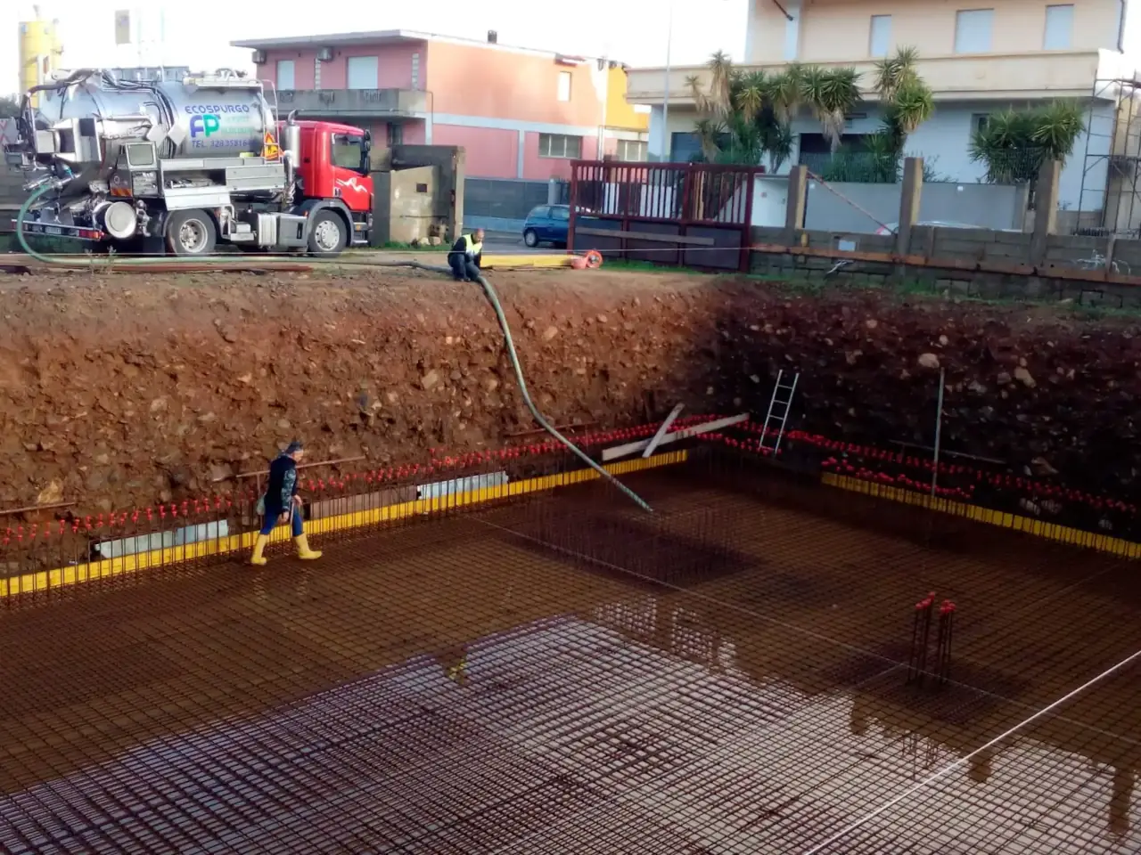 Mezzo autospurgo posizionato sul bordo di uno scavo di fondazione mentre un tubo flessibile raggiunge la zona di lavoro sottostante, con operatori impegnati nelle operazioni di aspirazione in un cantiere edile.