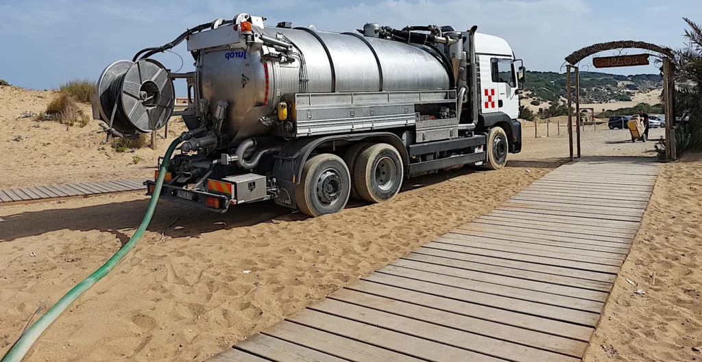 Mezzo autospurgo con cisterna metallica parcheggiato su una spiaggia, collegato con un tubo verde vicino a una passerella in legno.