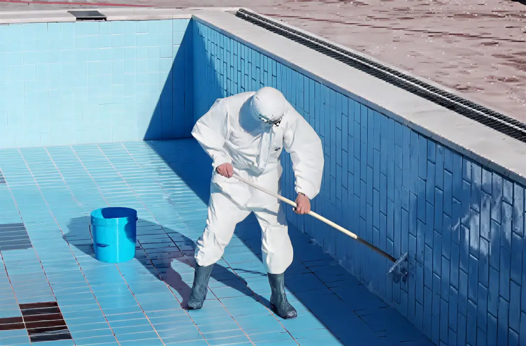 Operatore in tuta protettiva che pulisce l’interno di una piscina vuota con un attrezzo a manico lungo.